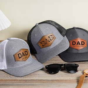 May include: Three gray trucker hats with leather patches that read "DAD". One hat has "EST. 2023" on the patch. Sunglasses and a watch with a brown leather band are also on the wooden surface.