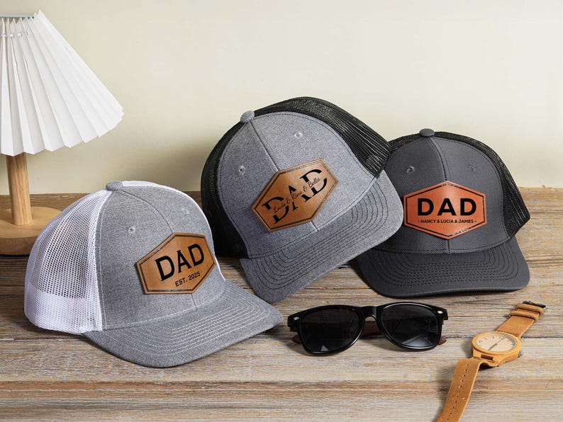 May include: Three gray trucker hats with leather patches that read "DAD". One hat has "EST. 2023" on the patch. Sunglasses and a watch with a brown leather band are also on the wooden surface.
