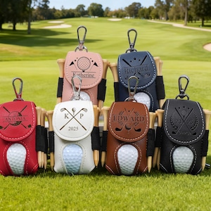 K&ouml;nnte beinhalten: Eine Auswahl an personalisierten Golfballhaltern in verschiedenen Farben, darunter Rot, Rosa, Wei&szlig;, Marineblau, Braun und Schwarz. Jeder Halter enth&auml;lt einen Golfball, Tees und einen Namen oder Initialen. Der Hintergrund ist ein gr&uuml;ner Golfplatz.