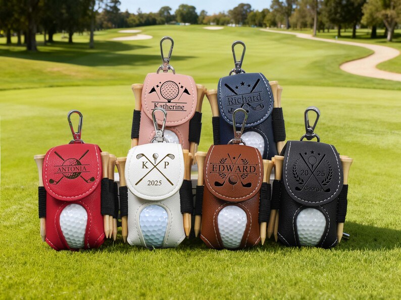 K&ouml;nnte beinhalten: Eine Auswahl an personalisierten Golfballhaltern in verschiedenen Farben, darunter Rot, Rosa, Wei&szlig;, Marineblau, Braun und Schwarz. Jeder Halter enth&auml;lt einen Golfball, Tees und einen Namen oder Initialen. Der Hintergrund ist ein gr&uuml;ner Golfplatz.