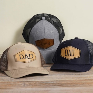 May include: Three baseball caps with mesh backs and leather patches. One cap is tan with a brown mesh, another is navy blue with a black mesh, and the third is gray with a black mesh. Each patch has the word "DAD" or "Dad" in a bold font.