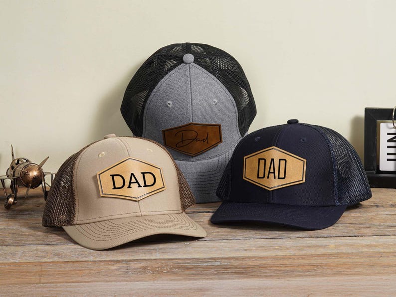 May include: Three baseball caps with mesh backs and leather patches. One cap is tan with a brown mesh, another is navy blue with a black mesh, and the third is gray with a black mesh. Each patch has the word "DAD" or "Dad" in a bold font.