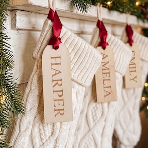 May include: Three cream-colored cable knit stockings hanging in a row. Each stocking features a wooden tag tied with a dark red satin ribbon. The tags are engraved with the names Harper, Amelia, and Emily in a clear, capitalized serif font.