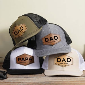 Può includere: Quattro cappellini da baseball di diversi colori e design. I cappellini hanno una rete sul retro e una toppa in pelle sul davanti. Le toppe hanno le parole "DAD" e "PAPA" con date diverse.