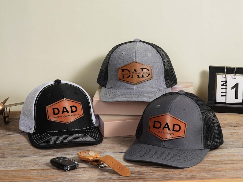 May include: Three baseball caps with mesh backs and leather patches. The patches read "DAD" in bold letters. One cap is black and white, another is gray, and the third is dark gray. A watch, car keys, and a calendar are also visible.