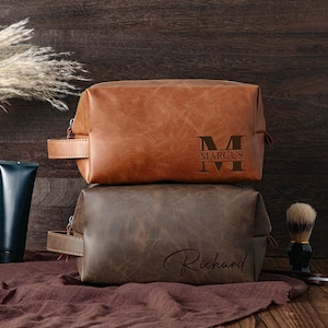 May include: Two leather toiletry bags, one tan and one brown, stacked on top of each other. The top bag has "Marcus" engraved on it. The bottom bag has "Richard" engraved on it. A dark blue tube and shaving brush are in the background.