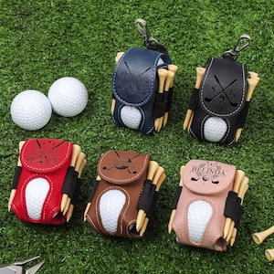 K&ouml;nnte beinhalten: Personalisierte Golfball- und Teehalter-Sets in Marineblau, Schwarz, Rot, Braun und Rosa. Jeder Halter enth&auml;lt einen Golfball und Holz-Tees. Die Halter sind mit Namen und Golfmotiven personalisiert.