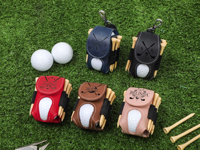 K&ouml;nnte beinhalten: Personalisierte Golfball- und Teehalter-Sets in Marineblau, Schwarz, Rot, Braun und Rosa. Jeder Halter enth&auml;lt einen Golfball und Holz-Tees. Die Halter sind mit Namen und Golfmotiven personalisiert.