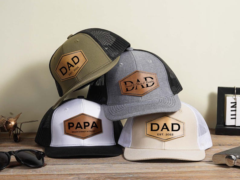 May include: A collection of trucker hats with personalized leather patches. The hats are in various colors, including olive green, gray, white, and tan. Each patch features the word "DAD" or "PAPA" in bold letters, with additional text and dates.