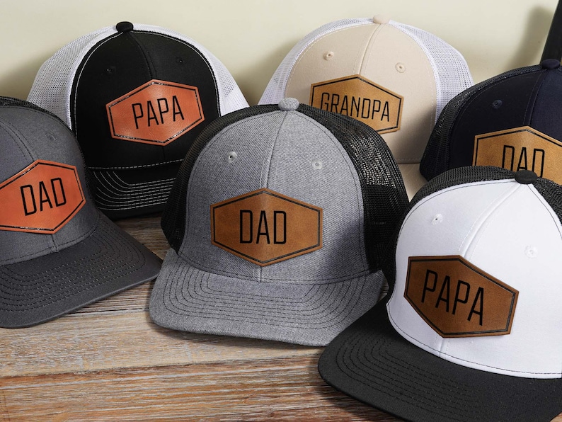 May include: A collection of trucker hats with mesh backs and various color combinations. Each hat features a leather patch with the words "DAD," "PAPA," or "GRANDPA" in black lettering. The hats are displayed on a wooden surface.