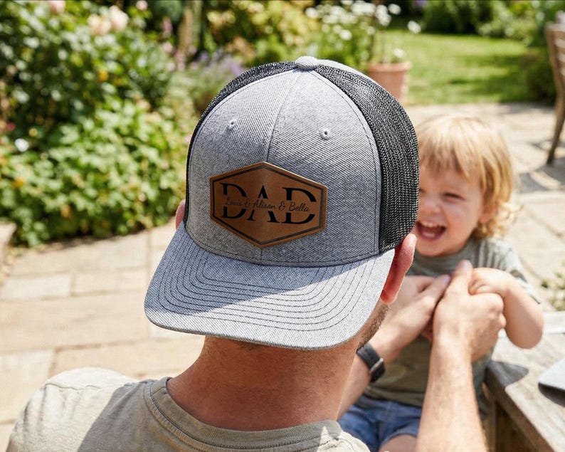 May include: Gray and black trucker hat with a brown leather patch that reads "DAD" with the names "Liam & Bella" below. The hat has a curved brim and a mesh back. The hat is being worn outdoors.