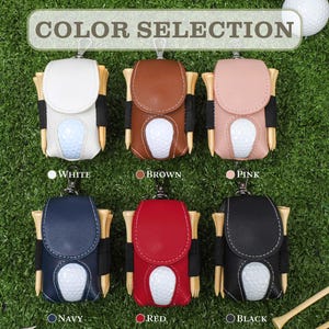 K&ouml;nnte beinhalten: Auswahl an Golfballhaltern in Wei&szlig;, Braun, Rosa, Marineblau, Rot und Schwarz. Jeder Halter hat einen Golfball und Holztees. Der Text "COLOR SELECTION" steht oben, mit Farbbezeichnungen darunter.