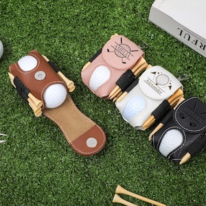 K&ouml;nnte beinhalten: Eine Sammlung personalisierter Golfballhalter aus braunem, rosa, wei&szlig;em und schwarzem Leder. Jeder Halter hat einen Golfball und Holz-Tees. Die Halter sind mit Namen und Golfmotiven personalisiert. Die Artikel sind auf einer gr&uuml;nen Oberfl&auml;che mit Golfb&auml;llen und einem Divot-Reparaturwerkzeug angeordnet.