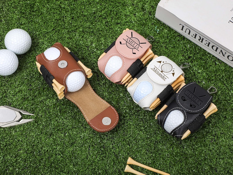 K&ouml;nnte beinhalten: Eine Sammlung personalisierter Golfballhalter aus braunem, rosa, wei&szlig;em und schwarzem Leder. Jeder Halter hat einen Golfball und Holz-Tees. Die Halter sind mit Namen und Golfmotiven personalisiert. Die Artikel sind auf einer gr&uuml;nen Oberfl&auml;che mit Golfb&auml;llen und einem Divot-Reparaturwerkzeug angeordnet.
