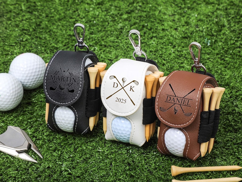 K&ouml;nnte beinhalten: Drei personalisierte Golfballhalter aus schwarzem, wei&szlig;em und braunem Leder, jeweils mit einem silbernen Clip. Die Halter enthalten Golfb&auml;lle und Holz-Tees. Der schwarze Halter tr&auml;gt den Namen "MARTIN" und der braune Halter den Namen "DANIEL".