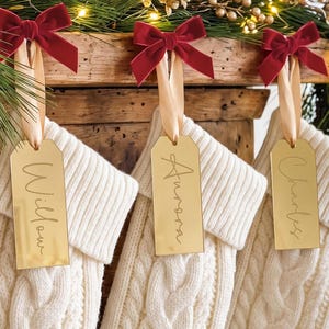 Può includere: Tre calze natalizie in maglia a trecce color crema con etichette rettangolari in acrilico dorato. Ogni etichetta è incisa con i nomi Willow, Aurora e Charles, fissate con un nastro color champagne e un fiocco in velluto rosso scuro.