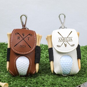 K&ouml;nnte beinhalten: Zwei personalisierte Golfballhalter mit Tees. Einer ist braun mit den Initialen "R T" und dem Jahr "2025", der andere ist wei&szlig; mit dem Namen "AMELIA". Beide haben einen silbernen Clip.