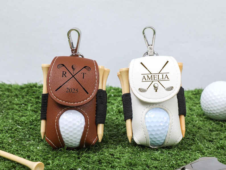 K&ouml;nnte beinhalten: Zwei personalisierte Golfballhalter mit Tees. Einer ist braun mit den Initialen "R T" und dem Jahr "2025", der andere ist wei&szlig; mit dem Namen "AMELIA". Beide haben einen silbernen Clip.