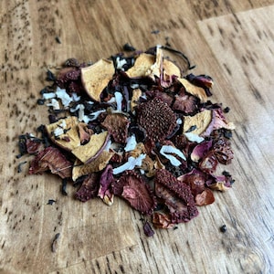 May include: A pile of loose leaf tea with dried fruit and flower pieces. The tea blend includes pieces of dried apple, strawberry, and coconut, with dark tea leaves and red flower petals. The tea is arranged on a wooden surface.