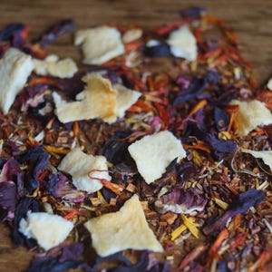 May include: A close-up of a blend of loose leaf tea with dried fruit pieces, hibiscus flowers, and other botanicals. The tea blend is a mix of red, orange, and purple hues.