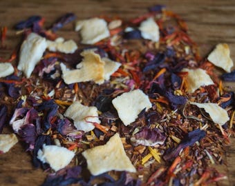 May include: A close-up of a blend of loose leaf tea with dried fruit pieces, hibiscus flowers, and other botanicals. The tea blend is a mix of red, orange, and purple hues.