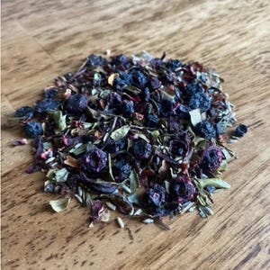 May include: A close-up of a blend of dried herbs and blueberries, including lavender, rose petals, and green tea leaves. The mixture is a light brown color with dark blue blueberries scattered throughout.