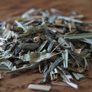 May include: A close-up of a pile of dried herbs and leaves, including green tea, lemongrass, and other botanicals. The herbs are loose and scattered on a wooden surface.