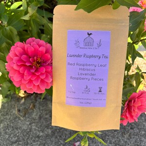 May include: A brown paper pouch of Lavender Raspberry Tea with a light purple label. The label features a rooster logo, ingredients including red raspberry leaf, hibiscus, lavender, and raspberry pieces, and brewing instructions for 1 oz (28g).