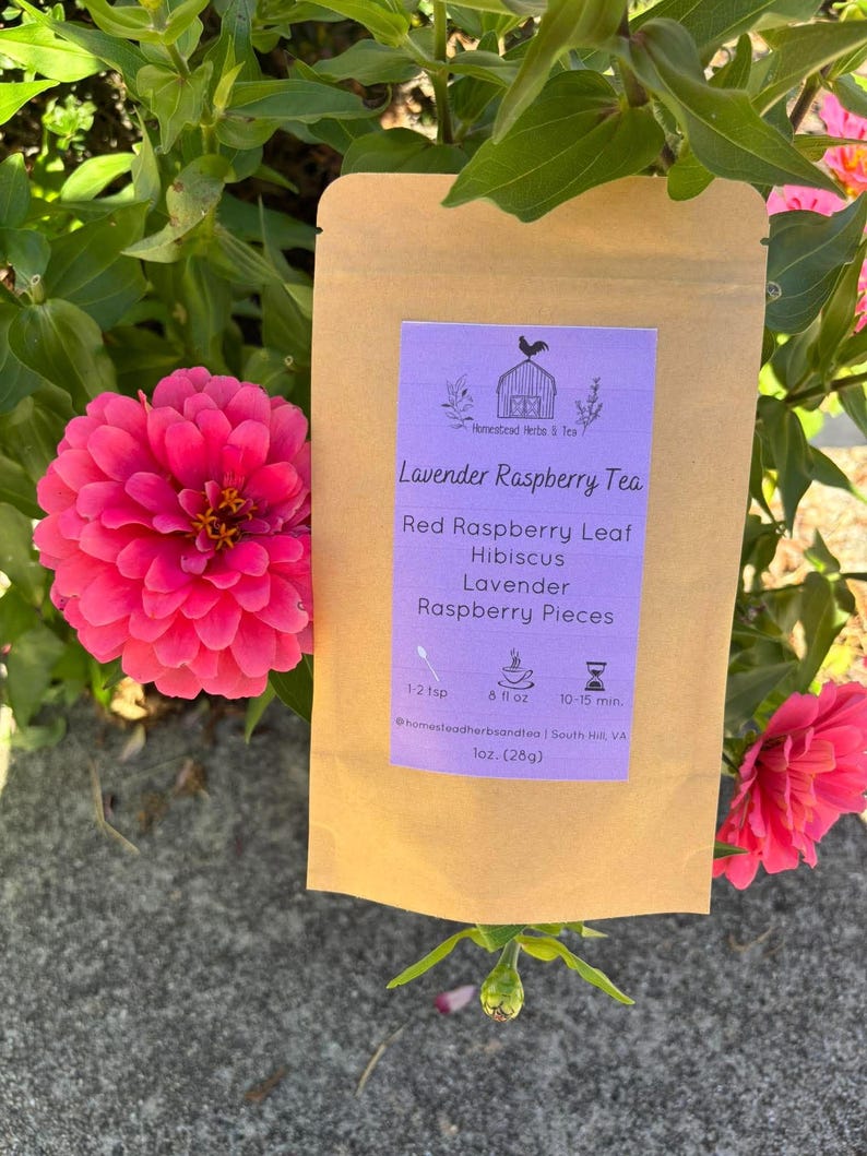 May include: A brown paper pouch of Lavender Raspberry Tea with a light purple label. The label features a rooster logo, ingredients including red raspberry leaf, hibiscus, lavender, and raspberry pieces, and brewing instructions for 1 oz (28g).