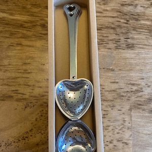 Op de afbeelding: Een hartvormige thee-ei van zilverkleurig metaal, met een lange steel en een scharnierend ontwerp. De thee-ei wordt gepresenteerd in een rechthoekige kartonnen doos, ideaal om cadeau te geven. Het heeft kleine gaatjes om losse thee te zetten.
