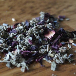 May include: A loose leaf herbal tea blend consisting of dried purple lavender buds, deep red hibiscus petals, and textured green sage leaves. The mixture is composed of small, fragmented botanical pieces with varied shapes and earthy tones.