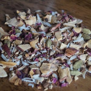 May include: A close-up of a blend of dried fruit, herbs, and spices for tea. The mix includes dried apple, coconut, rose petals, and green cardamom pods.