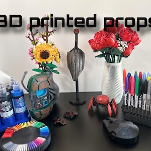 May include: A collection of 3D printed props, including a sunflower and rose bouquet, a robot, a spider, a spaceship, and a marker holder. The props are displayed on a black surface with paint bottles and a color wheel.