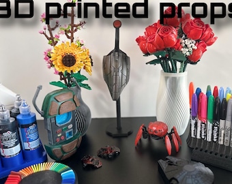 May include: A collection of 3D printed props, including a sunflower and rose bouquet, a robot, a spider, a spaceship, and a marker holder. The props are displayed on a black surface with paint bottles and a colour wheel.