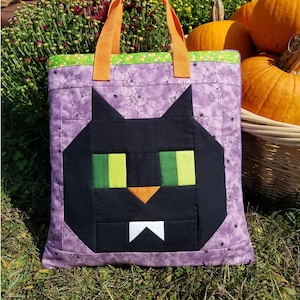 May include: A purple tote bag with a black cat face design. The cat has green eyes and a white mustache. The bag has orange handles and a green and white trim. The bag is sitting on a bed of green grass. The text "sew fun" is in a red circle with a green star. The text "Black Cat Tote Bag" is in black text. The text "www.sewfun.com" is in black text. The text "#122" is in black text.
