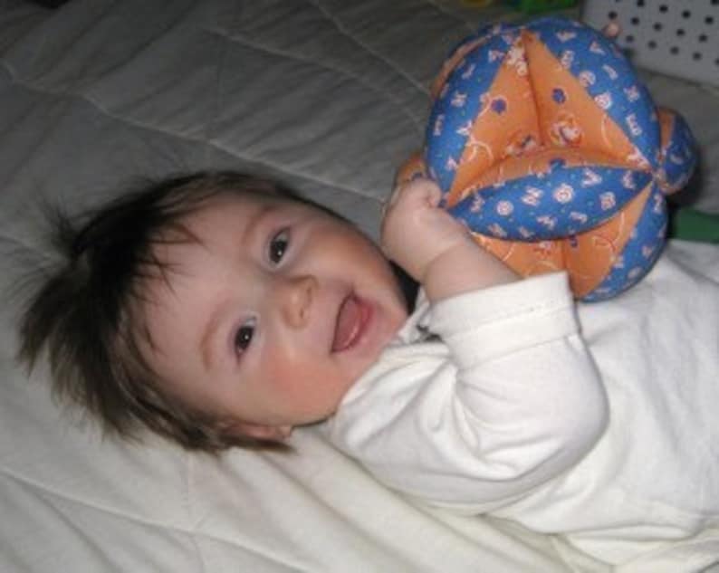 Puede incluir: Un beb&eacute; acostado sobre una manta blanca, sonriendo y sosteniendo una pelota de tela azul y naranja.
