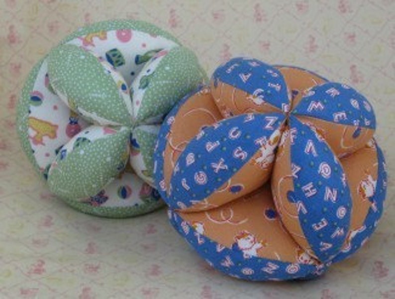 Puede incluir: Dos bolas de tela, una verde y otra azul, con un dise&ntilde;o estampado blanco y naranja. Las bolas est&aacute;n hechas de m&uacute;ltiples paneles de tela cosidos juntos.