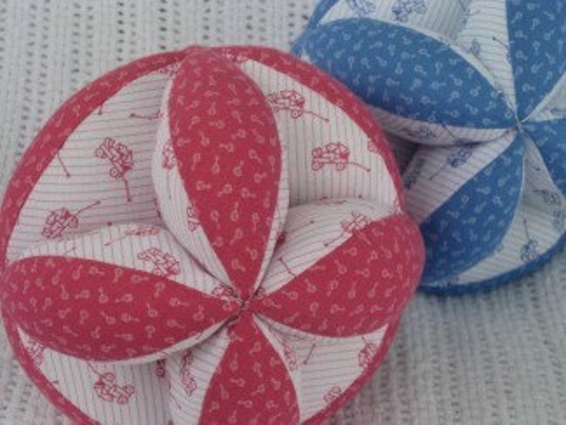 Puede incluir: Dos pelotas de tela para beb&eacute;s, una roja con rayas blancas y un peque&ntilde;o estampado de coche, y otra azul con lunares blancos.