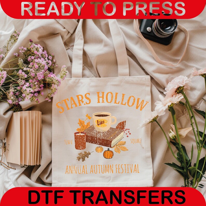 May include: A cream-colored tote bag with the words "Stars Hollow" in orange lettering. The bag features an illustration of a coffee cup, book, candle, and pumpkins. The words "Annual Autumn Festival" are printed below the illustration.
