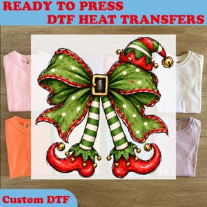May include: A DTF heat transfer design featuring a large green bow with white polka dots, a black belt buckle, and an elf hat with a gold bell. Below the bow are two striped green and white legs ending in curled red elf shoes with gold bells.