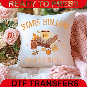 May include: White decorative pillow with an autumn-themed design. The pillow features the text "STARS HOLLOW" and "ANNUAL AUTUMN FESTIVAL". The design includes a coffee cup, book, candle, pumpkins, and fall leaves.