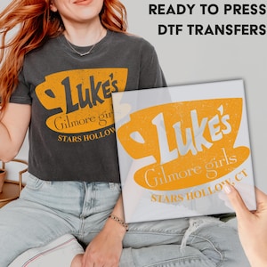 May include: A white heat transfer design with a yellow coffee cup and the text "Luke's Gilmore Girls STARS HOLLOW, CT".