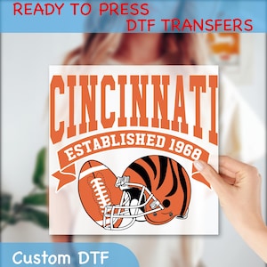 May include: A white DTF transfer featuring the word "CINCINNATI" in orange block letters, with "ESTABLISHED 1968" below. Includes an orange football and helmet with black stripes. Text at the top reads "READY TO PRESS DTF TRANSFERS" and "Custom DTF" at the bottom.