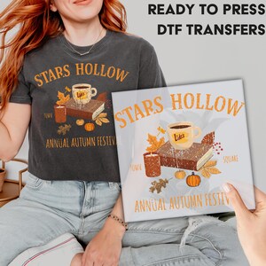 May include: A charcoal gray t-shirt and a square print featuring the text "STARS HOLLOW" in orange. The design includes a coffee cup, a book, autumn leaves, a candle, and pumpkins. The text "ANNUAL AUTUMN FESTIVAL" is also present.
