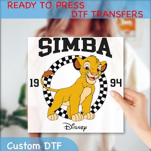 May include: A white iron-on transfer with a cartoon image of Simba from Disney's The Lion King. Simba is standing in a checkered circle with the text "SIMBA" above him and "19 94" on either side of the circle. The text "DISNEY" is below the circle.