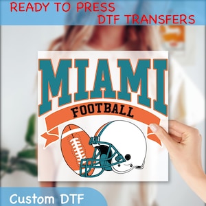 May include: A white square featuring a teal and orange design with the word "MIAMI" above "FOOTBALL". Below the text are illustrations of a football and a helmet. The words "READY TO PRESS DTF TRANSFERS" and "Custom DTF" are also present.