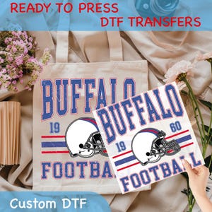 May include: A beige tote bag and a white transfer sheet featuring a distressed design with the text "BUFFALO FOOTBALL" in blue and white, along with a football helmet graphic. The text "READY TO PRESS DTF TRANSFERS" is at the top.