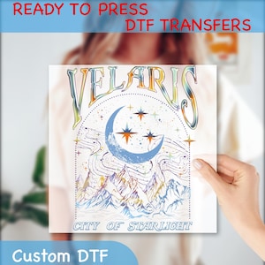 May include: A ready-to-press DTF transfer featuring a colorful graphic design of a crescent moon, stars, and mountains with the text "Velaris City of Starlight".