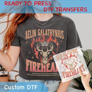May include: A black t-shirt with a graphic design featuring a stag with antlers, a crown, and flames. The text "Aelin Galathynius Fireheart" is printed on the shirt. The text "She was fire, and light, and ash, and embers. She was Aelin Fireheart, and she bowed for no one and nothing" is printed below the graphic.