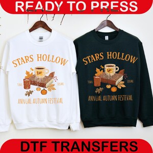 May include: White and dark green sweatshirts with the text "STARS HOLLOW" in orange. Each sweatshirt features a graphic of a coffee cup, book, candles, and pumpkins. The text "ANNUAL AUTUMN FESTIVAL" is below the graphic. The words "READY TO PRESS" and "DTF TRANSFERS" are at the top and bottom.